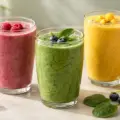 Tre glass med rosa, grønn og gul smoothie omgitt av friske bringebær, blåbær, mango og spinat.